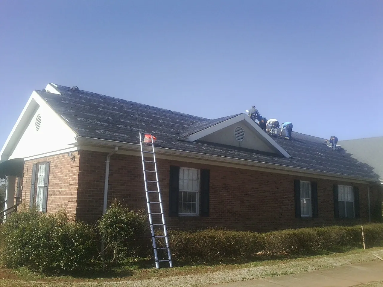 Crew tearing shingles off a brick ranch roof with a ladder set — Emergency Roof Repair work by our crew in Fairmount