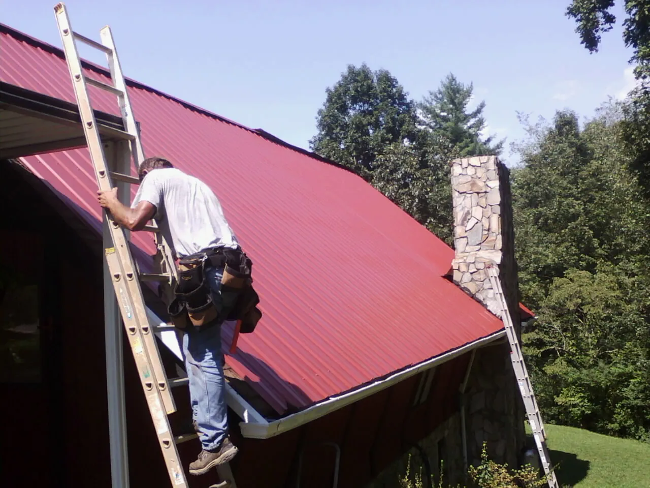 Roofer on a ladder beside a red metal roof and stone chimney — Soffit & Fascia Repair by Roofing Brothers in Fairmount